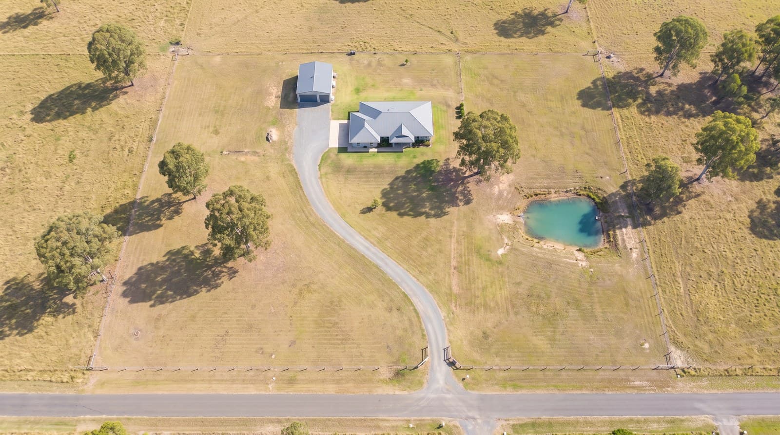 Aerial view of an acreage block with a long driveway, paddocks and a farm dam.