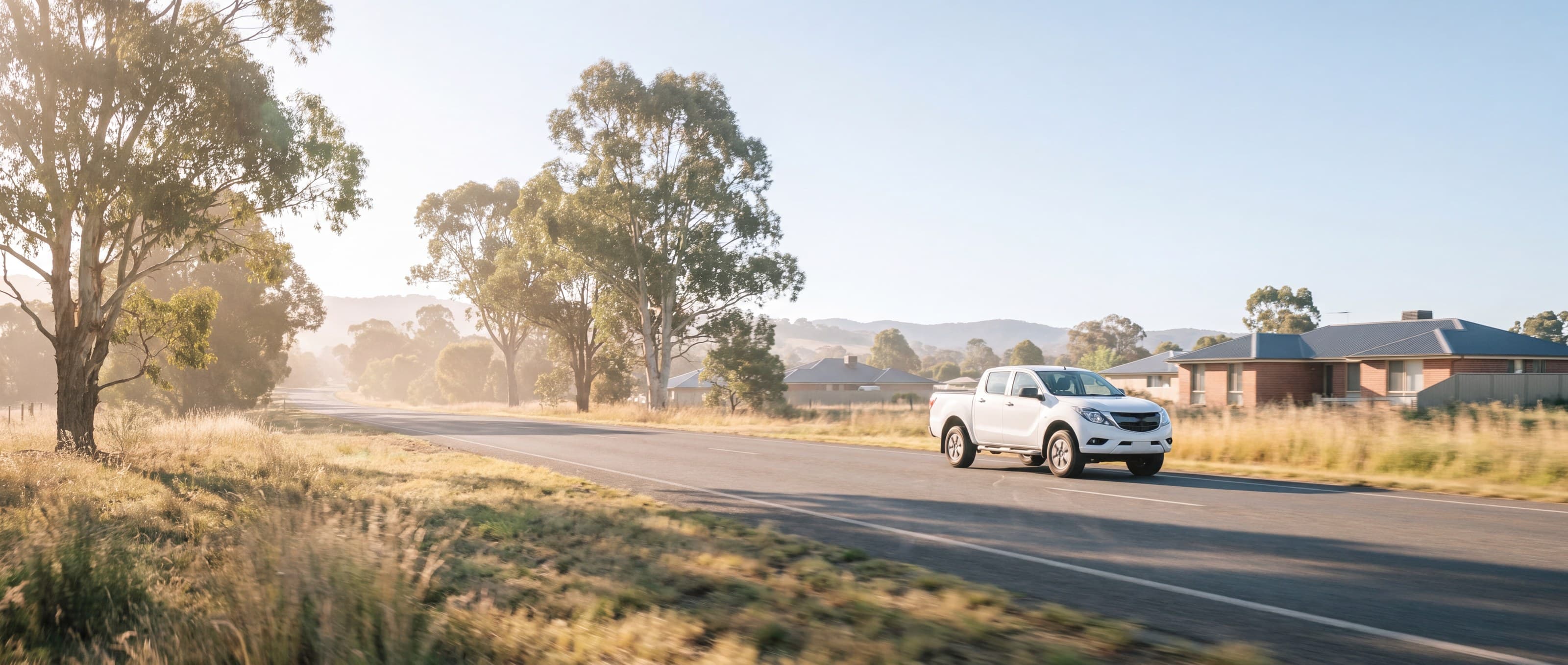 A white ute driving along a quiet country road in early morning light.